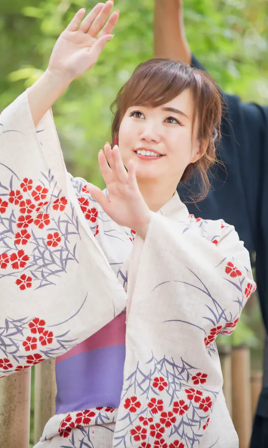 Bon Odori A woman in a floral yukata raises her arms gracefully while smiling outdoors, with greenery and bamboo fencing behind her. A man in traditional attire stands in the background, also raising his arms.