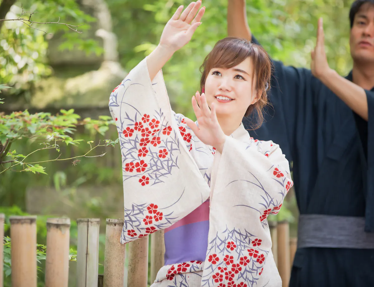 Bon Odori A woman in a floral yukata raises her arms gracefully while smiling outdoors, with greenery and bamboo fencing behind her. A man in traditional attire stands in the background, also raising his arms.