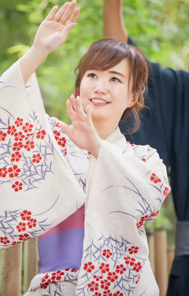 Bon Odori A woman in a floral yukata raises her arms gracefully while smiling outdoors, with greenery and bamboo fencing behind her. A man in traditional attire stands in the background, also raising his arms.