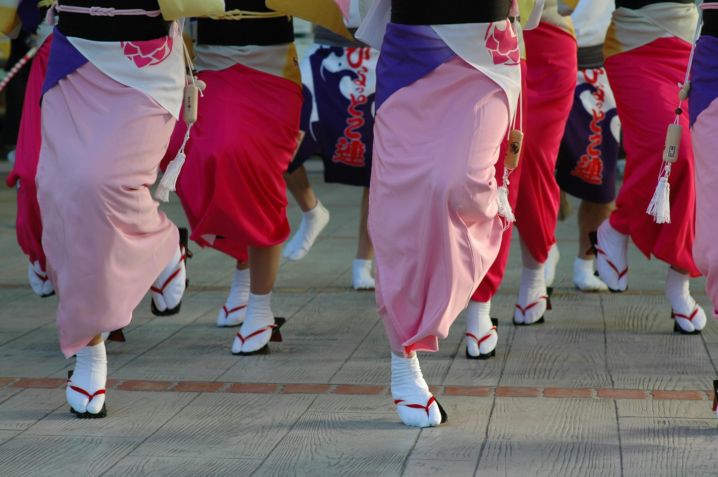 A group of people in colorful traditional Japanese attire perform a dance, wearing pink and red kimono skirts, white socks, and wooden sandals on a paved surface.