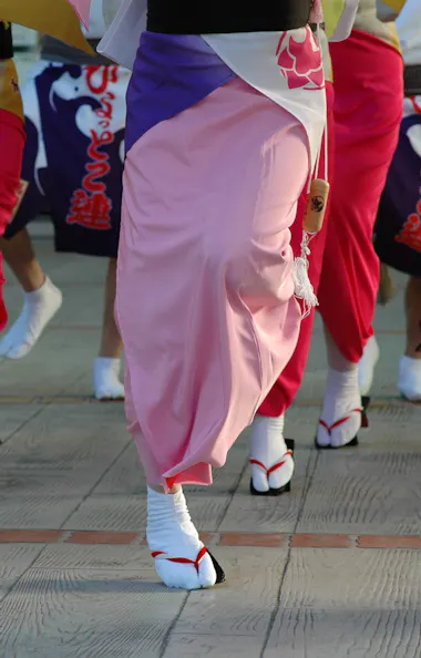 Bon Odori A group of people in colorful traditional Japanese attire perform a dance, wearing pink and red kimono skirts, white socks, and wooden sandals on a paved surface.