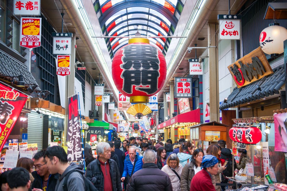 A busy indoor market in Japan with colorful signs, large red lanterns, and many people shopping and walking through the crowded aisles beneath a bright, covered ceiling.