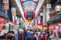 A busy indoor market in Japan with colorful signs, large red lanterns, and many people shopping and walking through the crowded aisles beneath a bright, covered ceiling.