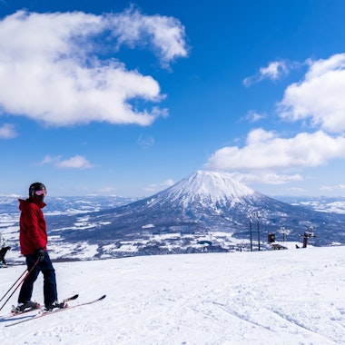 Skiing A skier in a red jacket stands on a snowy slope with ski poles, while others ski nearby. In the background, a snow-covered mountain rises under a blue sky with scattered clouds.