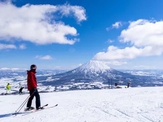 A skier in a red jacket stands on a snowy slope with ski poles, while others ski nearby. In the background, a snow-covered mountain rises under a blue sky with scattered clouds.
