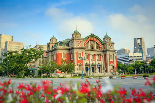 A historic red and beige building with domes and arched windows stands in a city park, surrounded by greenery and red flowers, with modern skyscrapers in the background under a blue sky.