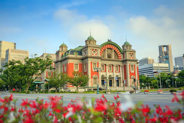 A historic red and beige building with domes and arched windows stands in a city park, surrounded by greenery and red flowers, with modern skyscrapers in the background under a blue sky.