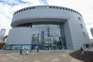 A modern, circular glass and metal building with people walking in front. The sign at the top is in Japanese, and the tall building is surrounded by a paved plaza under a partly cloudy sky.