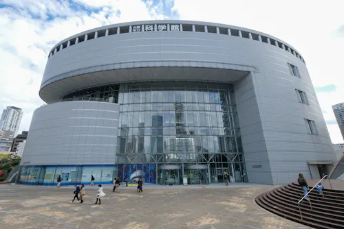 A modern, circular glass and metal building with people walking in front. The sign at the top is in Japanese, and the tall building is surrounded by a paved plaza under a partly cloudy sky.