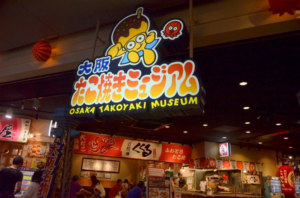 A colorful sign for the Osaka Takoyaki Museum hangs above a busy food court, featuring a cartoon octopus holding takoyaki and surrounded by bright, playful lettering in Japanese and English.