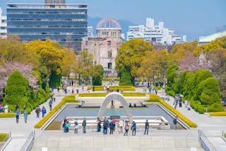 People visiting the Hiroshima Peace Memorial Park in spring. The image features the cenotaph, the reflecting pool, and the A-Bomb Dome in the background, with trees and buildings surrounding the area on a clear day.