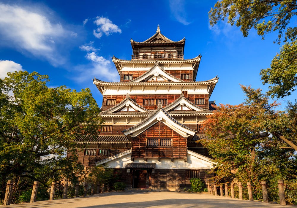 A traditional Japanese castle with multiple tiered roofs stands surrounded by trees under a bright blue sky with scattered clouds. Sunlight highlights the wooden and white exterior of the building.