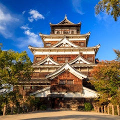 Hiroshima Castle A traditional Japanese castle with multiple tiered roofs stands surrounded by trees under a bright blue sky with scattered clouds. Sunlight highlights the wooden and white exterior of the building.