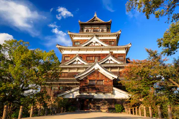 Hiroshima Castle A traditional Japanese castle with multiple tiered roofs stands surrounded by trees under a bright blue sky with scattered clouds. Sunlight highlights the wooden and white exterior of the building.