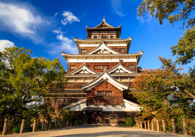 A traditional Japanese castle with multiple tiered roofs stands surrounded by trees under a bright blue sky with scattered clouds. Sunlight highlights the wooden and white exterior of the building.