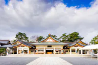 Traditional Japanese shrine with ornate, curved roofs and wooden details, set against a dramatic sky with clouds. Trees frame the building, and a stone path leads to the entrance.