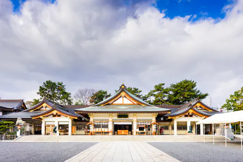 Traditional Japanese shrine with ornate, curved roofs and wooden details, set against a dramatic sky with clouds. Trees frame the building, and a stone path leads to the entrance.