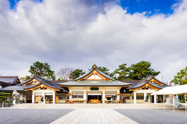 Traditional Japanese shrine with ornate, curved roofs and wooden details, set against a dramatic sky with clouds. Trees frame the building, and a stone path leads to the entrance.