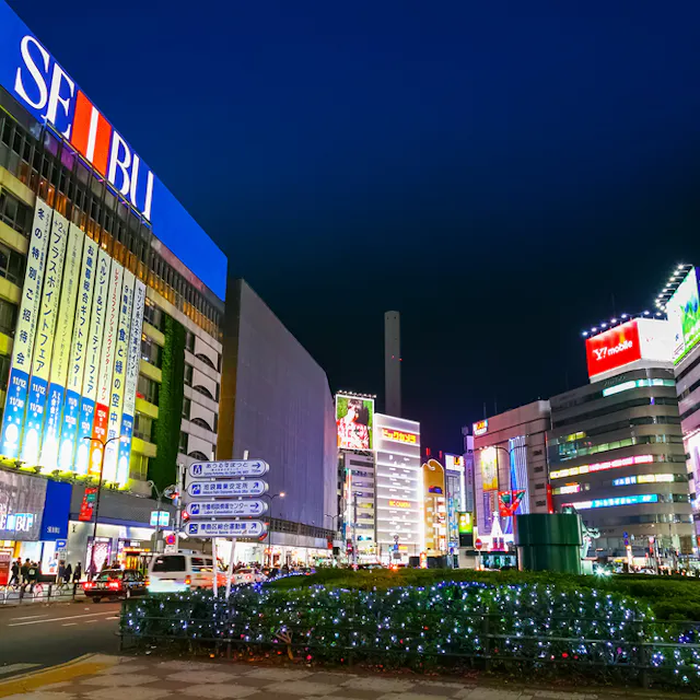 Ikebukuro A vibrant city street at night in Japan, illuminated by colorful neon signs and advertisements on tall buildings, with cars, people, and festive string lights on greenery in the foreground.