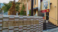 Entrance of the Kyoto International Manga Museum with a sign in Japanese and English, nearby trees, stairs, and a colorful poster featuring manga artwork on the building wall.