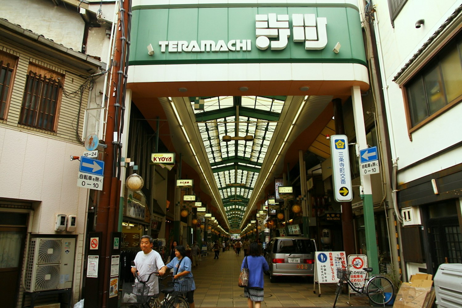 Entrance to Teramachi shopping arcade in Kyoto, Japan. People walk and cycle under a covered walkway lined with shops. Signs with Japanese text hang above, and colorful lanterns are visible inside.