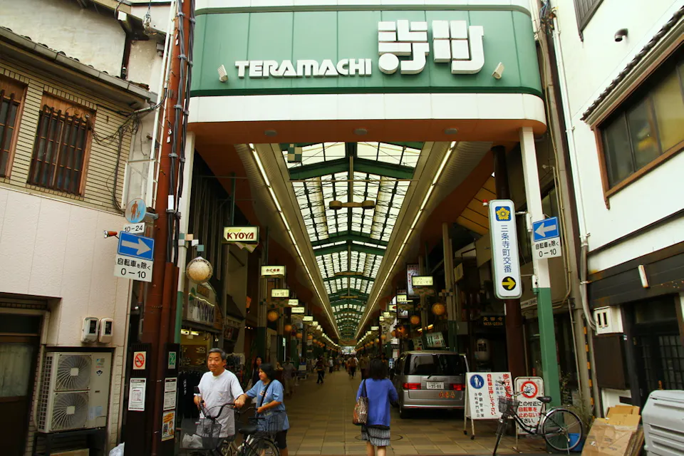 Teramachi and Shinkyogoku Shopping Arcades Entrance to Teramachi shopping arcade in Kyoto, Japan. People walk and cycle under a covered walkway lined with shops. Signs with Japanese text hang above, and colorful lanterns are visible inside.