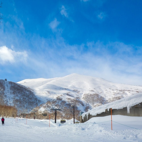 Niseko Annupuri International Ski Area A person skis on a snowy slope near a ski lift and a building with icicles, with snow-covered mountains and a bright blue sky in the background.