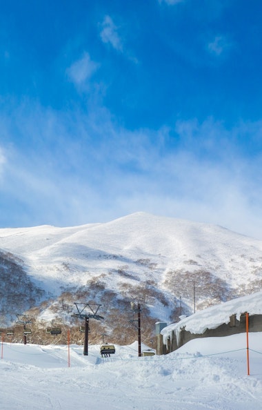 Niseko Annupuri International Ski Area A person skis on a snowy slope near a ski lift and a building with icicles, with snow-covered mountains and a bright blue sky in the background.