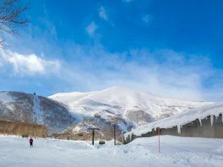A person skis on a snowy slope near a ski lift and a building with icicles, with snow-covered mountains and a bright blue sky in the background.