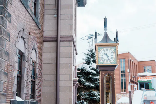 A vintage street clock with a gold frame stands outside in a snowy urban area, surrounded by brick buildings and snow-covered trees on a cloudy winter day.