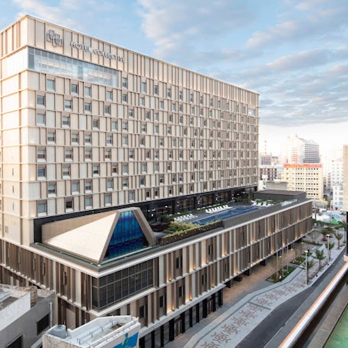 Hotel Collective An aerial view of a large, modern hospital building with multiple stories and a sleek, rectangular design. The hospital features a rooftop garden area with plants and seating. Surrounding the hospital are other urban buildings and a partly cloudy sky in the background.