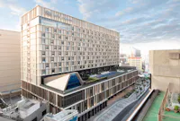 An aerial view of a large, modern hospital building with multiple stories and a sleek, rectangular design. The hospital features a rooftop garden area with plants and seating. Surrounding the hospital are other urban buildings and a partly cloudy sky in the background.