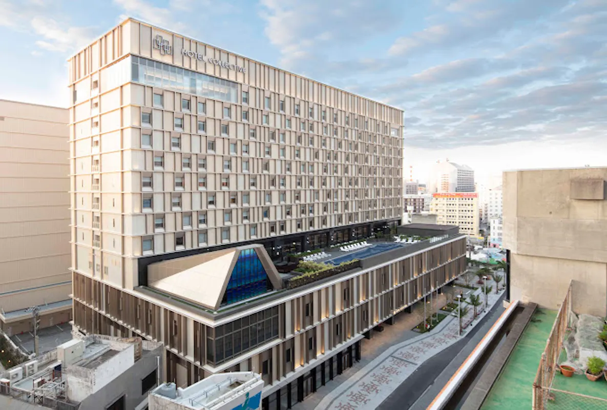 An aerial view of a large, modern hospital building with multiple stories and a sleek, rectangular design. The hospital features a rooftop garden area with plants and seating. Surrounding the hospital are other urban buildings and a partly cloudy sky in the background.