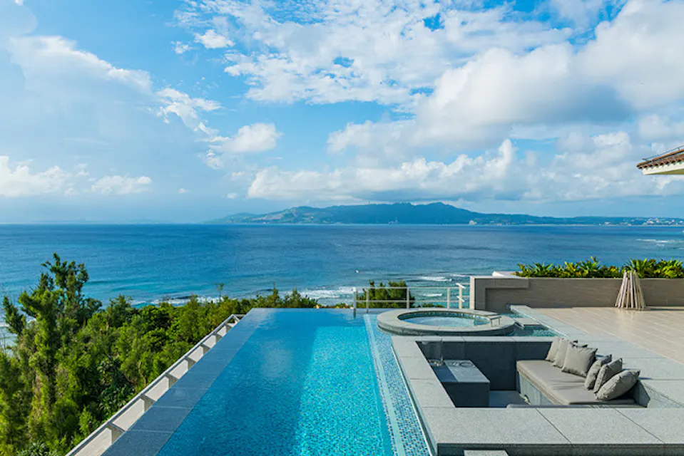 Halekulani Okinawa A luxurious infinity pool overlooks a vast blue ocean with a stunning coastal view. The pool area includes a sunken seating area and a hot tub. Bright, fluffy clouds dot the blue sky above, and lush greenery surrounds the space, creating an idyllic setting.