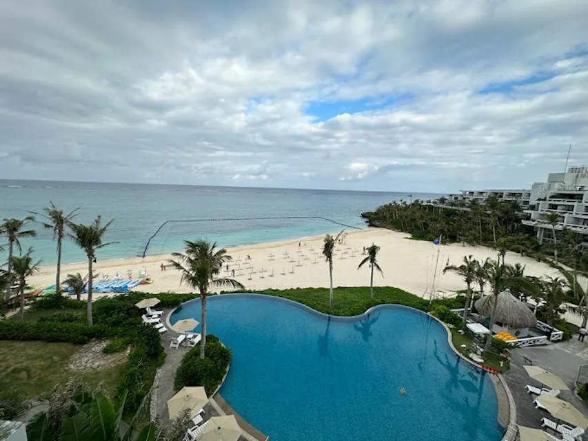 Hotel Moon Beach A view from above of a resort featuring a large, curvy pool with lounge chairs and palm trees. Beyond the pool, a white sandy beach meets a calm blue ocean, with some light clouds in the sky. Several beachfront buildings are visible to the right.