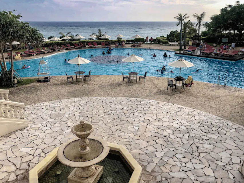 Hotel Nikko Alivila A scenic view of a resort's outdoor swimming pool surrounded by lounge chairs and umbrellas. People are swimming and relaxing by the pool. Palm trees and the ocean are visible in the background, with a stone fountain in the foreground on a paved patio.