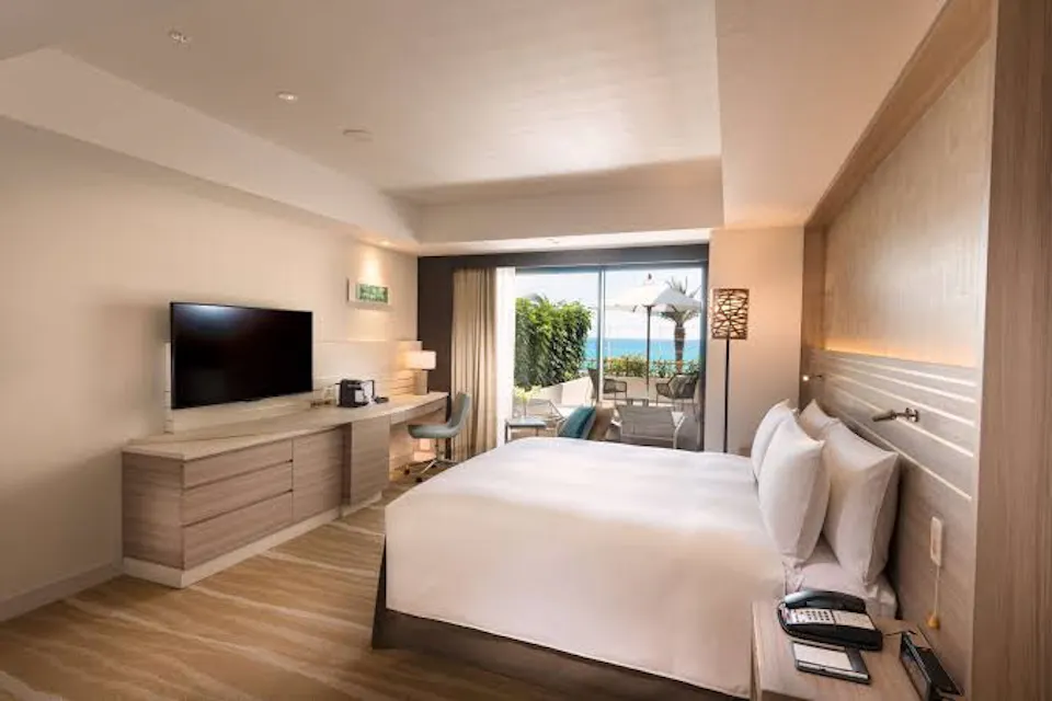 DoubleTree by Hilton Okinawa Chatan Resort A modern hotel room featuring a king-sized bed with white linens, a flat-screen TV on a wooden dresser, a coffee maker, a seating area, and a large window leading to a balcony with a view of the ocean. The room is well-lit with contemporary decor.