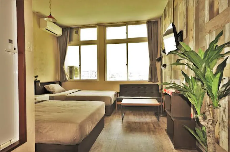 My Place A cozy room with two single beds, a small sofa, a wall-mounted TV, a coffee table, and a large indoor plant. The room has wooden decor and large windows with curtains, letting in ample natural light. An air conditioner is mounted above one of the beds.