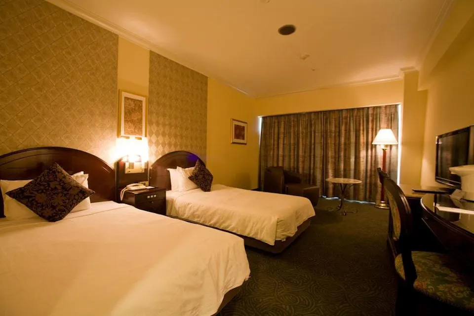 Okinawa Kariyushi Urban Resort Naha A hotel room with two neatly made single beds, each with a decorative pillow. The room features a patterned accent wall, a nightstand with a lamp and telephone, a TV on a desk, a seating area with a floor lamp and round table, and a window with heavy curtains.