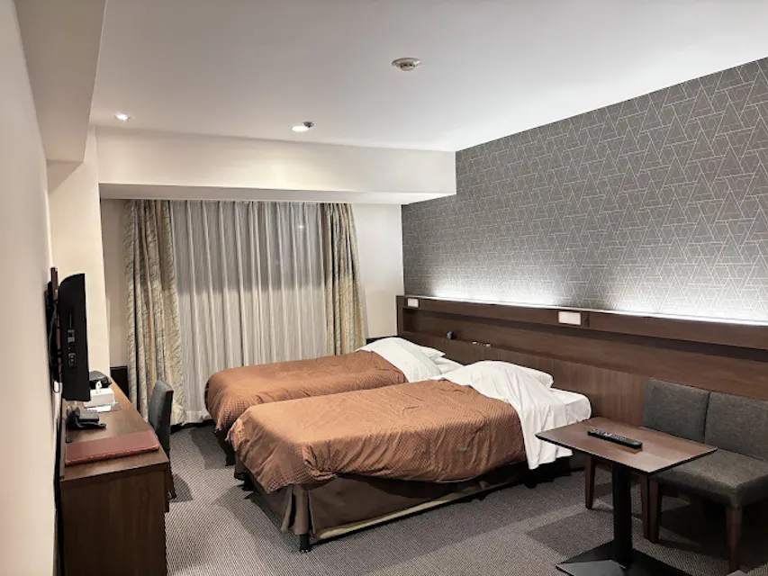 Hotel Sun Okinawa A modern hotel room featuring two single beds with brown bedspreads, a gray sofa with a small table, a flat-screen TV mounted on a wall opposite to the beds, and a desk with a chair. The room is well-lit, and there is a large window with sheer and blackout curtains.