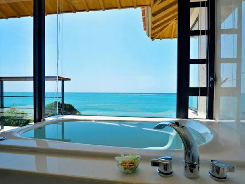 Hyakuna Garan A luxurious bath with a large tub situated by floor-to-ceiling windows overlooking a serene ocean view. The bath has modern fixtures and a small glass dish with amenities on the side. The bright blue sea contrasts with the clean, minimalistic design of the room.