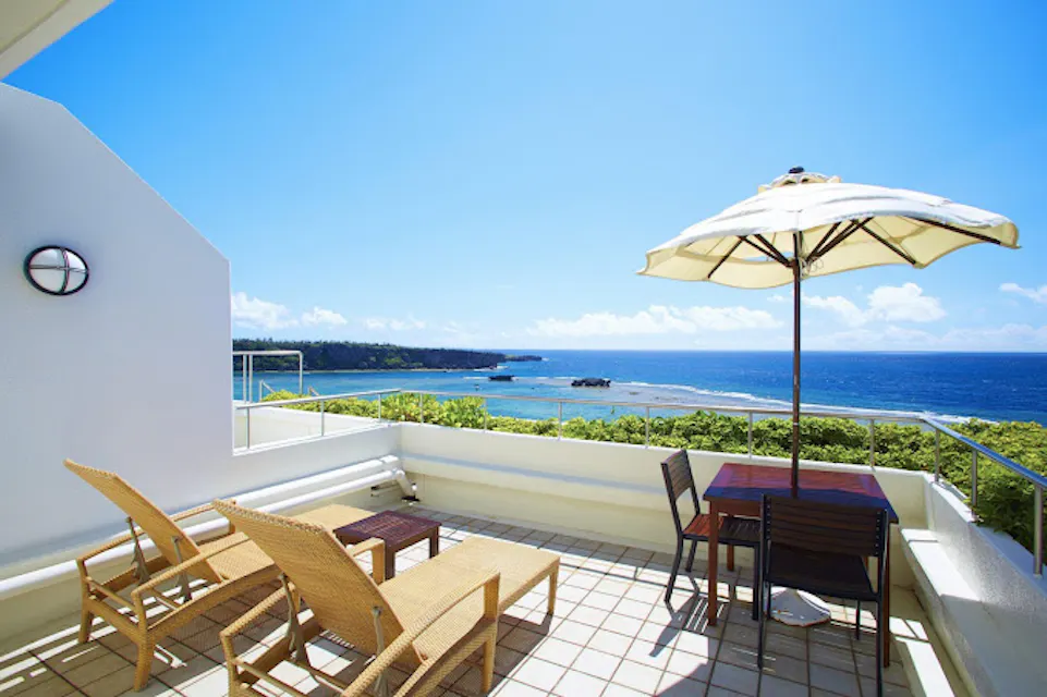 ANA InterContinental Manza Beach Resort A spacious patio with lounge chairs and a table with an umbrella overlooks a beautiful beach. The clear blue sky and ocean create a serene and picturesque view. Greenery borders the patio and leads to the sandy shore in the distance.