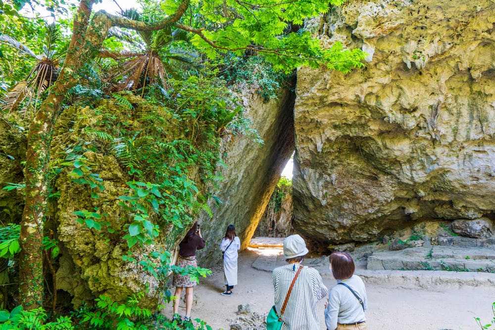 A group of people stands near large, mossy rock formations and lush green trees in a sunlit outdoor setting, observing the unique natural landscape and taking photos.