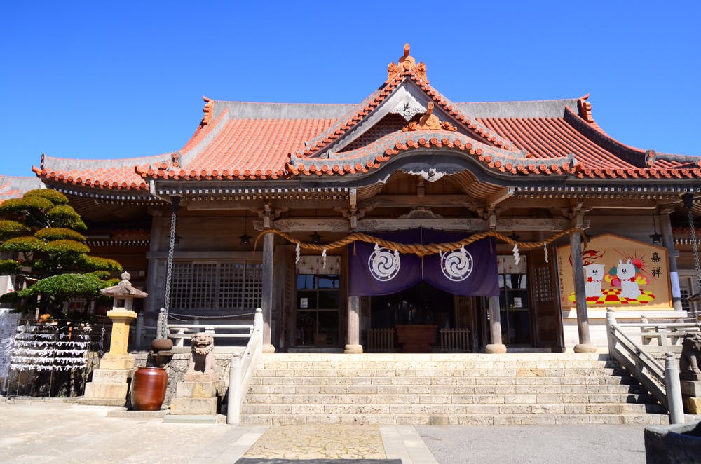 A traditional Japanese shrine with a red-tiled roof, stone steps, and purple banners. Sculpted trees, lanterns, and a decorative board are visible under a clear blue sky.