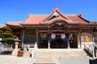 A traditional Japanese shrine with a red-tiled roof, stone steps, and purple banners. Sculpted trees, lanterns, and a decorative board are visible under a clear blue sky.