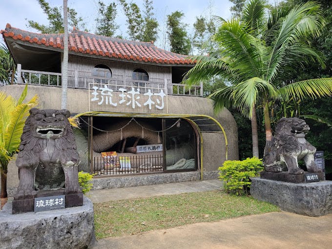 A building with a tiled roof, large sign with Japanese text, and two stone lion statues at the entrance, surrounded by tropical plants and trees.