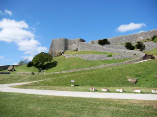 Stone walls of an ancient castle or fortress rise along a grassy hill under a blue sky with scattered clouds, with a winding path leading up the slope.