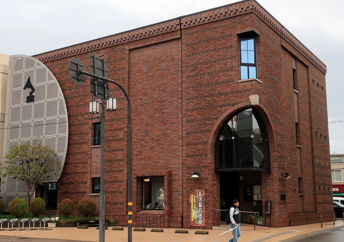A large, modern brick building with arched windows and geometric architectural features, including a circular metal structure on the left. A few people walk by on the street in front of the building.