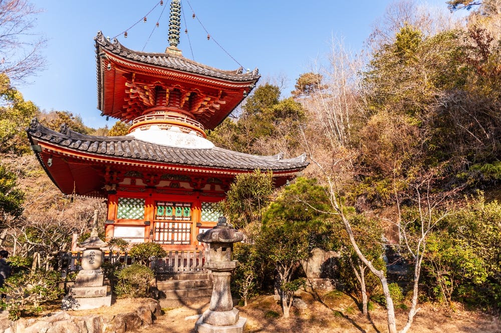 A traditional Japanese pagoda with ornate details stands among trees and shrubs on a sunny day, with a stone lantern in the foreground and a clear blue sky above.