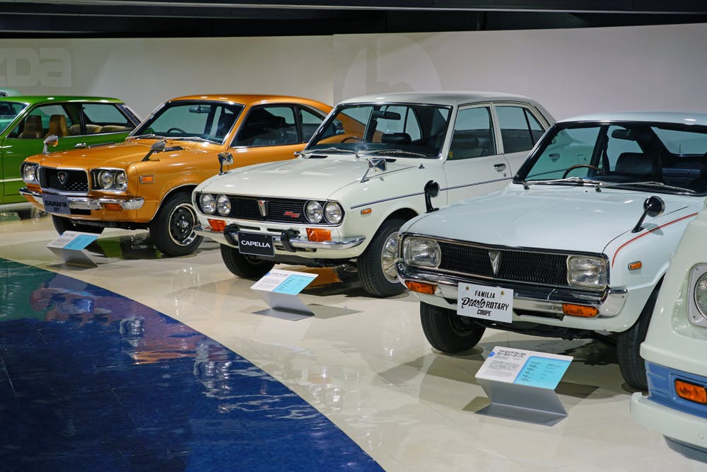 A row of vintage Mazda cars, including orange, white, and green models, is displayed in an indoor showroom, each with informational placards in front.